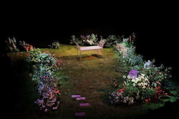 A stage set designed as a garden with green grass, colourful flowers, a white metal bench at the centre, and stepping stones leading towards it, all surrounded by darkness.