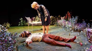 A woman stands in a garden setting, looking down with concern at a man lying on the grass. The man is wearing a patterned shirt and hat, whilst flowers and a bench surround them on stage.