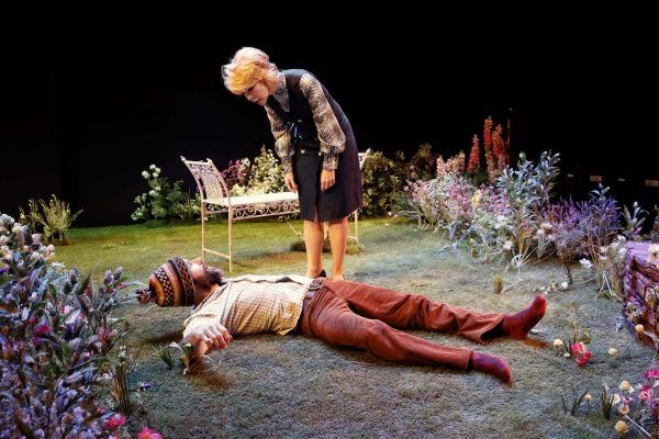 A woman stands in a garden setting, looking down with concern at a man lying on the grass. The man is wearing a patterned shirt and hat, whilst flowers and a bench surround them on stage.