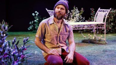 A bearded man in a knitted hat and short-sleeved shirt kneels on grass in a garden setting, surrounded by flowers, with a white metal bench and foliage in the background, under stage lighting.