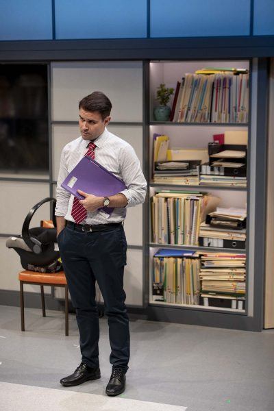 A man in office attire stands holding a purple folder, looking thoughtful. Behind him are shelves filled with neatly stacked and upright files and paperwork. A chair and bag are also visible.