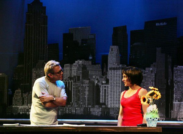 A man in a grey shirt and glasses talks to a woman in a red sleeveless top beside a table with a vase of sunflowers, with a city skyline backdrop behind them.
