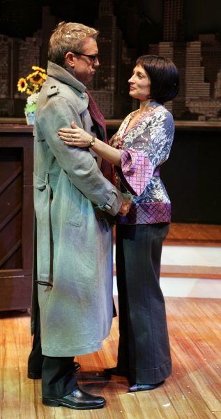 A man in a grey trench coat and scarf stands facing a woman with a short dark bob. She wears a patterned blouse and dark trousers. They hold hands and look at each other on a stage with a flower vase in the background.