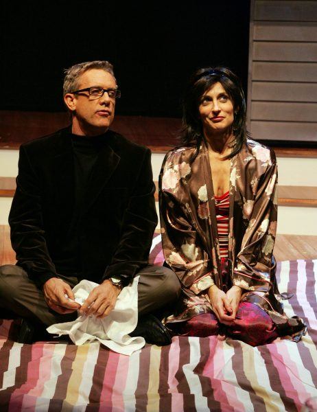 A man and a woman sit side by side on a colourful striped blanket on stage. The man wears glasses, a black jacket, and grey trousers, while the woman wears a floral dressing gown over a striped top, both looking slightly to the side.