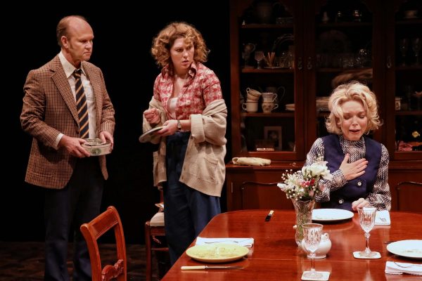 Three actors perform a dramatic scene in a dining room; a man in a checked jacket and tie stands holding a dish, a woman in a floral blouse looks concerned, and an older woman sits at the table with her hand on her chest.