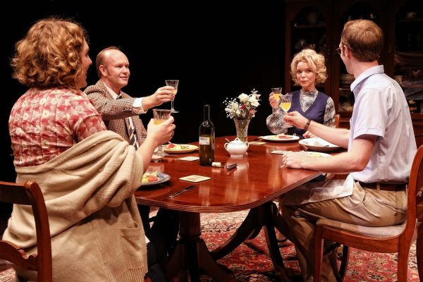 Four adults sit around a wooden dining table, raising glasses in a toast. The table is laid with plates of food, a wine bottle, and a flower vase. They appear to be engaged in a formal dinner gathering.
