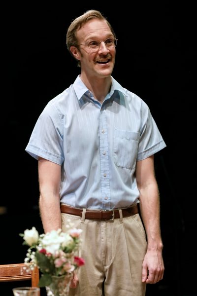 A man with glasses and a moustache, wearing a light blue short-sleeve button-up shirt and beige trousers, stands and smiles next to a table with a small bouquet of flowers. The background is dark.