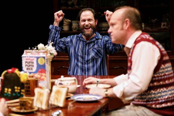 Two men sit at a breakfast table. One man in a striped shirt smiles broadly with raised fists, celebrating. The other man, wearing a jumper vest, looks away pensively. Toast, cereal boxes, and pastries are on the table.