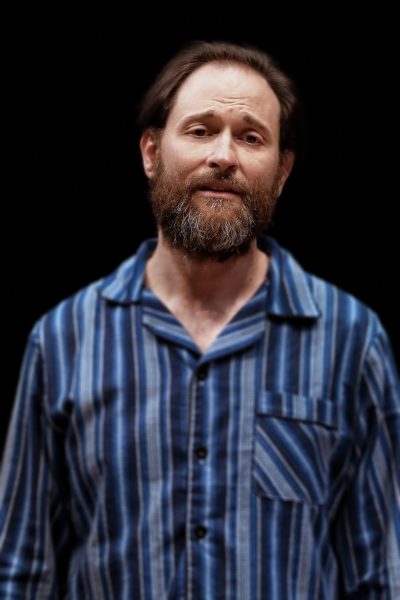 A man with a beard and moustache wearing a blue striped shirt stands against a black background, looking slightly up with a neutral or contemplative expression.