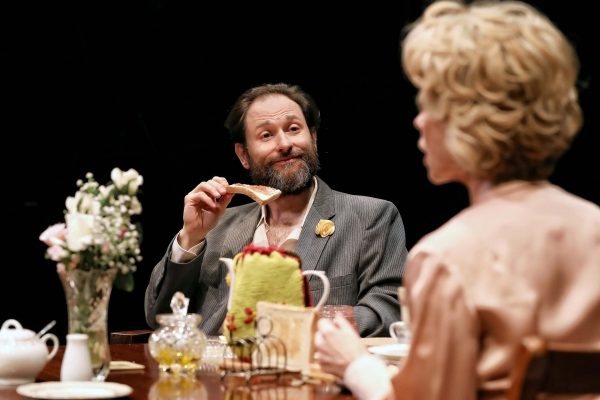 A bearded man in a suit, holding a piece of toast and smiling, sits at a table with a woman. The table is set with a teapot, cake, flowers, and various dishes; the scene appears to be from a stage play.