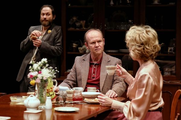 Three people sit around a dining table with teacups, plates, and a flower vase. One man stands holding food, another sits looking serious, and a woman in a satin dressing gown holds a teacup, facing away from the camera.