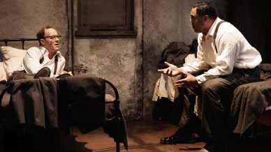 Two men talk intensely in a dimly lit, worn room; one sits up in bed while the other sits nearby, gesturing with both hands, both dressed in white shirts and dark trousers. The scene conveys tension and seriousness.