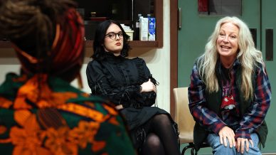 Three women sit in a room; one with long grey hair and a checked shirt smiles while speaking, another with black hair and glasses wears a black dress and sits with arms folded, and a third, seen from behind, wears a colourful patterned outfit.