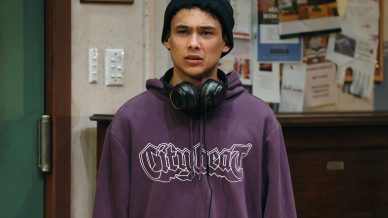 A young man wearing a black beanie, large headphones around his neck, and a purple hoodie with City Heat written on it stands indoors, looking slightly concerned. A noticeboard is visible in the background.