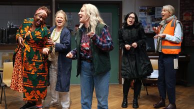 Five women stand together indoors, smiling and laughing. One wears a vibrant patterned dressing gown, another is in a waistcoat and flannel shirt, one is in a black dress, another in a high-vis vest, and one in light-coloured clothes.
