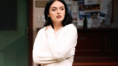 A woman with black hair wearing a white long-sleeved top stands indoors with her arms folded, looking to the side. A noticeboard and wooden furniture are visible in the background.