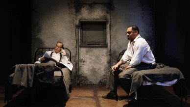 Two men sit on separate metal-frame beds in a dimly lit, grey-walled room. One man reclines, reading, while the other sits on the edge of his bed, looking thoughtful. The setting appears stark and sombre, resembling a prison cell.