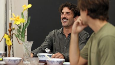 Two men sit at a dining table set with bowls and a vase of yellow flowers. One man, smiling and laughing, faces forwards while the other sits beside him, turned slightly away.
