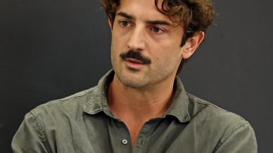 A man with curly brown hair and a moustache stands with arms crossed, wearing an olive green button-up shirt against a plain dark background.