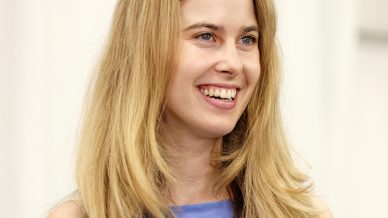 A young woman with long blonde hair smiles whilst wearing a sleeveless light purple top. The background is softly blurred and light-coloured.