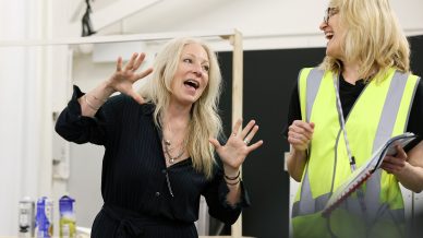 Two women are talking and laughing together. One, in a dark dress, gestures animatedly with her hands. The other, wearing glasses and a yellow safety vest, holds a clipboard and smiles. They seem to be enjoying a lively conversation indoors.
