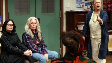 Four women are in a room; one older woman stands and speaks, gesturing with her hand, whilst two women sit and listen. The setting appears to be a community meeting or group discussion. Posters and a piano are visible in the background.