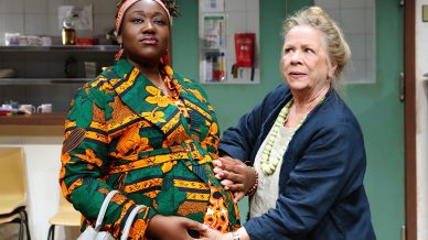 Two women stand indoors; one, wearing a colourful patterned dress and headscarf, is visibly pregnant and holding her bump, while the other, in a blue coat, gently touches her bump and looks supportive.