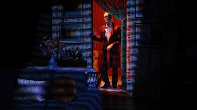 A person in a red dressing gown stands in a doorway beaded with strings, lit by a spotlight. The room features patterned wallpaper, a blue sofa with cushions, and a vase of flowers on a table, all in dim lighting.