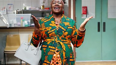 A woman in a colourful patterned dress stands indoors with her eyes closed and arms raised, holding a grey handbag. She appears expressive, possibly singing or speaking passionately, in a room with chairs and kitchen items behind her.