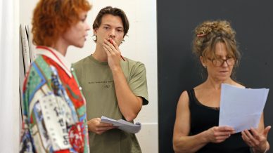 Three people stand indoors holding papers. One person in a colourful shirt is speaking, another in a green shirt listens thoughtfully with a hand on their face, and a third in a black top reads from a page.