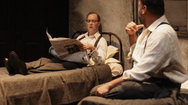Two men in dress shirts and braces sit on separate single beds in a dimly lit room. One man reads a newspaper while the other sits on the edge of his bed, gesturing as they appear to be engaged in conversation.