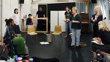 A group of people in a rehearsal space, some seated and some standing, engage in a discussion. Scripts and bags are on chairs and the floor. The atmosphere is collaborative and creative.