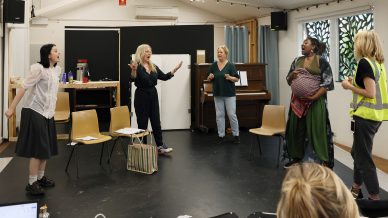 Five women rehearse energetically in a bright room; some are singing or gesturing passionately. Chairs and a piano are in the background, while one woman wears a reflective vest and another holds a patterned bundle.