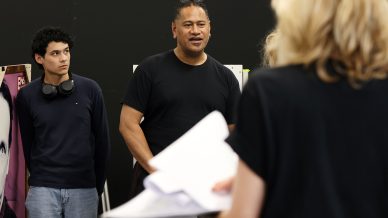 Three people in a rehearsal space: one man speaking in the centre, another man with headphones round his neck listens on the left, and a blonde person with papers faces away on the right.