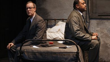 Two men in suits sit back-to-back on a metal bed in a dimly lit, sparse room with grey walls. Papers, a red box, and a belt are on the bed between them. Both appear deep in thought or emotion.