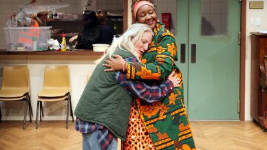Two women hug warmly in a room with green double doors and a kitchen area in the background. One woman has grey hair and wears casual clothes; the other wears bright, patterned traditional African attire and smiles.