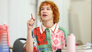 A woman with short, curly red hair and a colourful patterned shirt holds up a red pencil whilst speaking. She is seated at a table with books, a pink water bottle, and a pink flask in front of her.