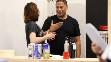 Two people stand and talk near a wooden counter with cartons, bottles, and a red mug. One person gestures with their hand while smiling; the other listens. A third person is partially visible, holding papers.