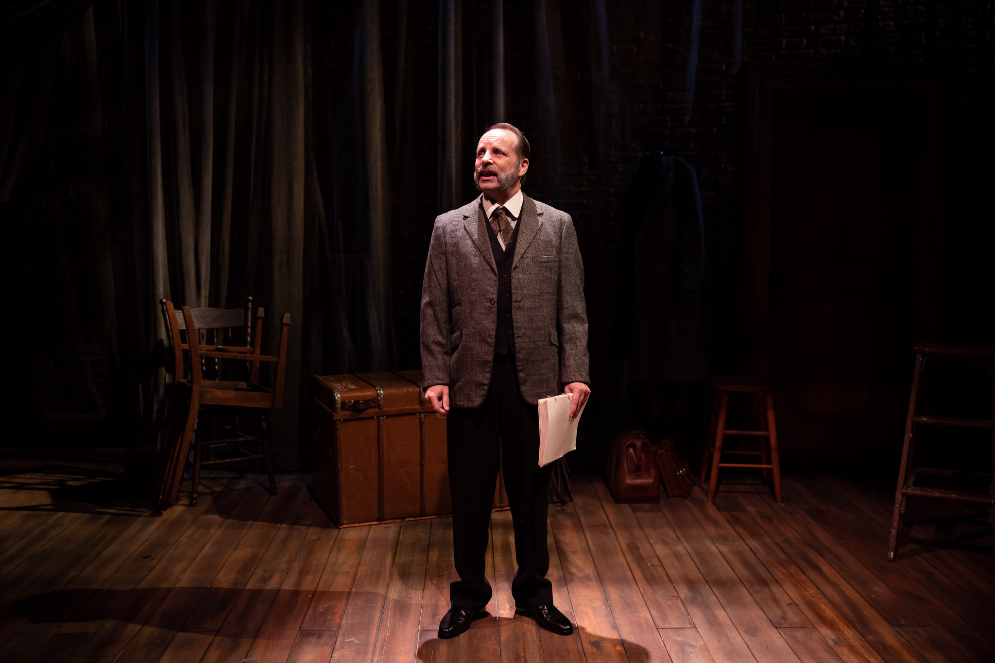 A man in a suit stands alone on a dimly lit stage, holding papers. Behind him are wooden chairs, a trunk, a coat on a hook, and dark curtains, creating a dramatic and sombre atmosphere.