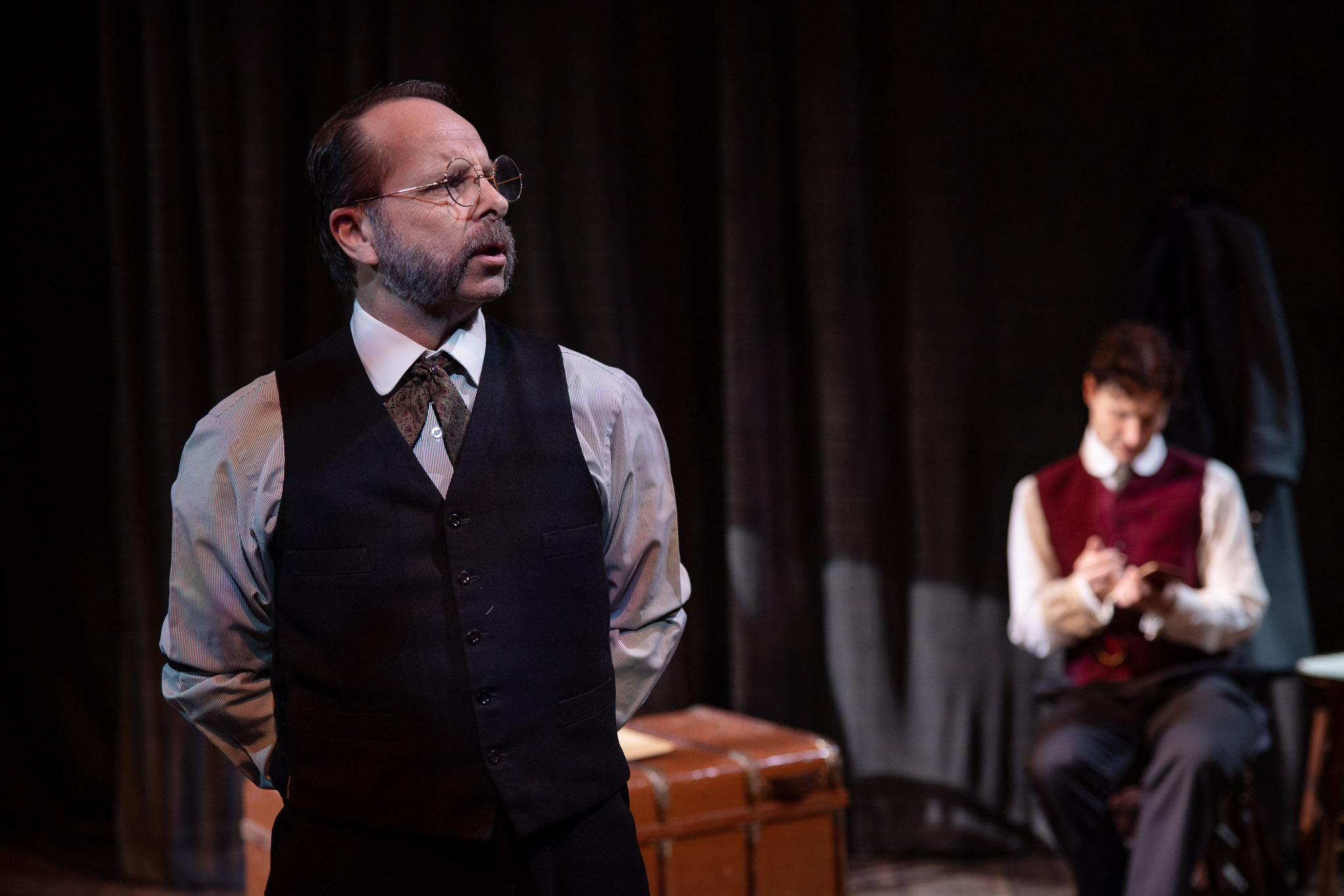 A man with glasses and a beard, wearing a waistcoat and tie, stands in the foreground on stage, speaking or singing. In the background, another man sits and writes, dressed in a red waistcoat. Dark curtains and a suitcase are visible.