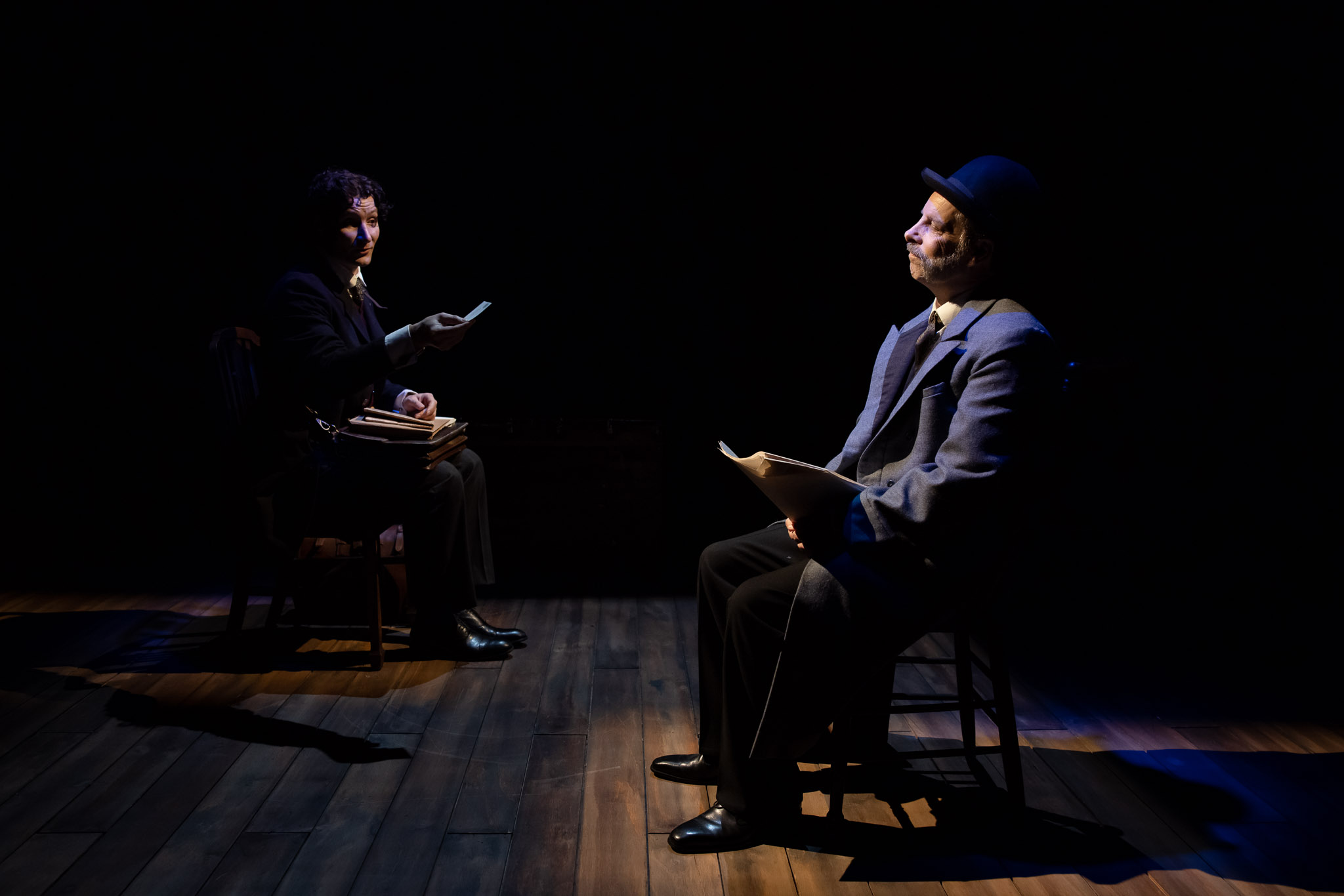 Two actors sit on stage facing each other, illuminated by spotlights against a dark background. One holds papers and wears a suit and hat; the other, also in a suit, appears to be reading or presenting a document.