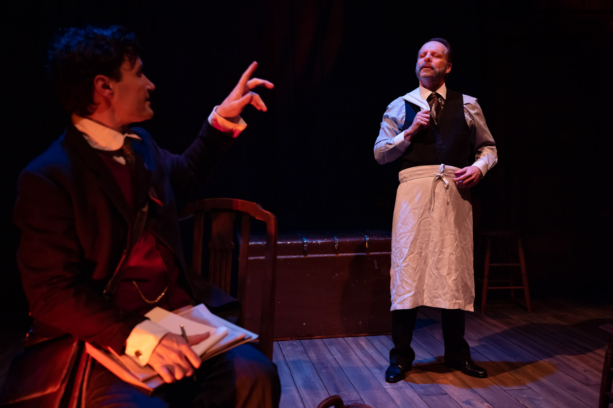 Two men on a dimly lit stage: one sits holding a notebook and pen, gesturing, while the other stands facing him, wearing a white apron over formal clothes, appearing to listen or respond.
