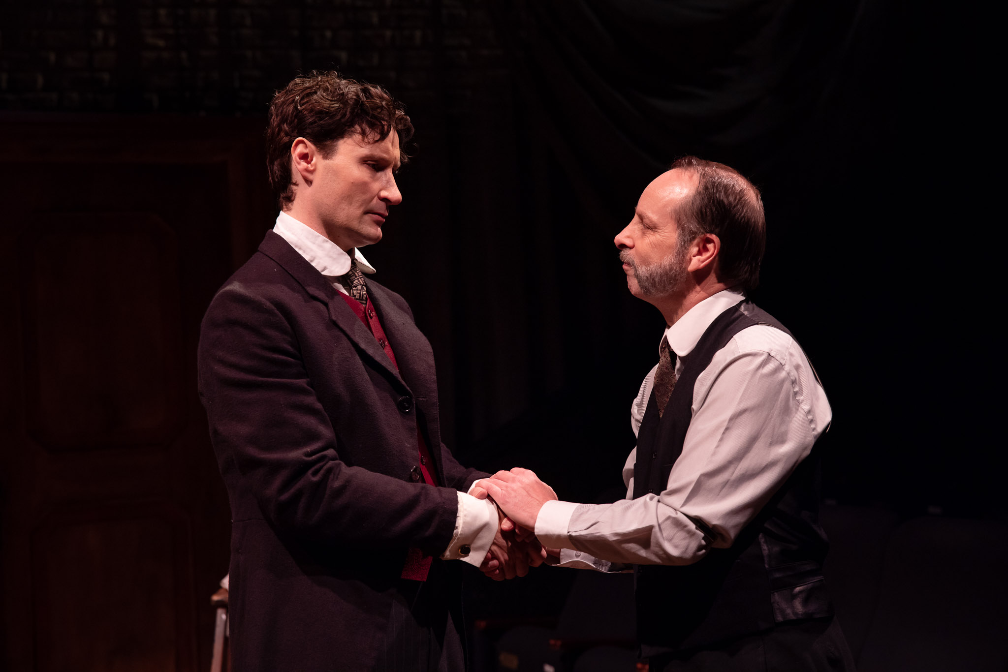 Two men in formal period clothing face each other on a dimly lit stage. One man grips the other's hands, and they share a serious, intense expression, suggesting an emotional or dramatic moment in a theatrical performance.