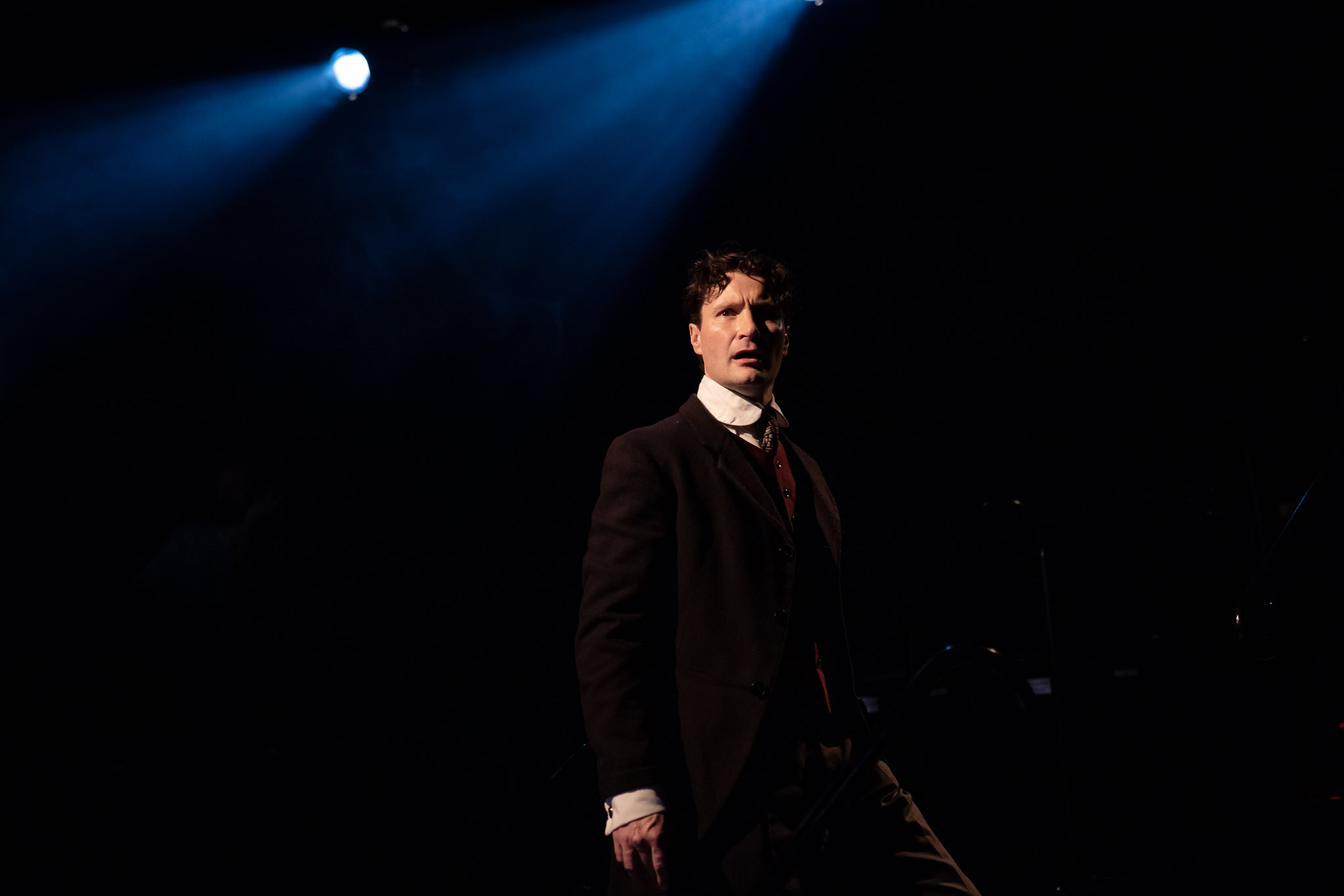 A man in a dark suit stands under a spotlight on a dimly lit stage, looking intently ahead. Blue stage lights shine from above, creating dramatic shadows around him.