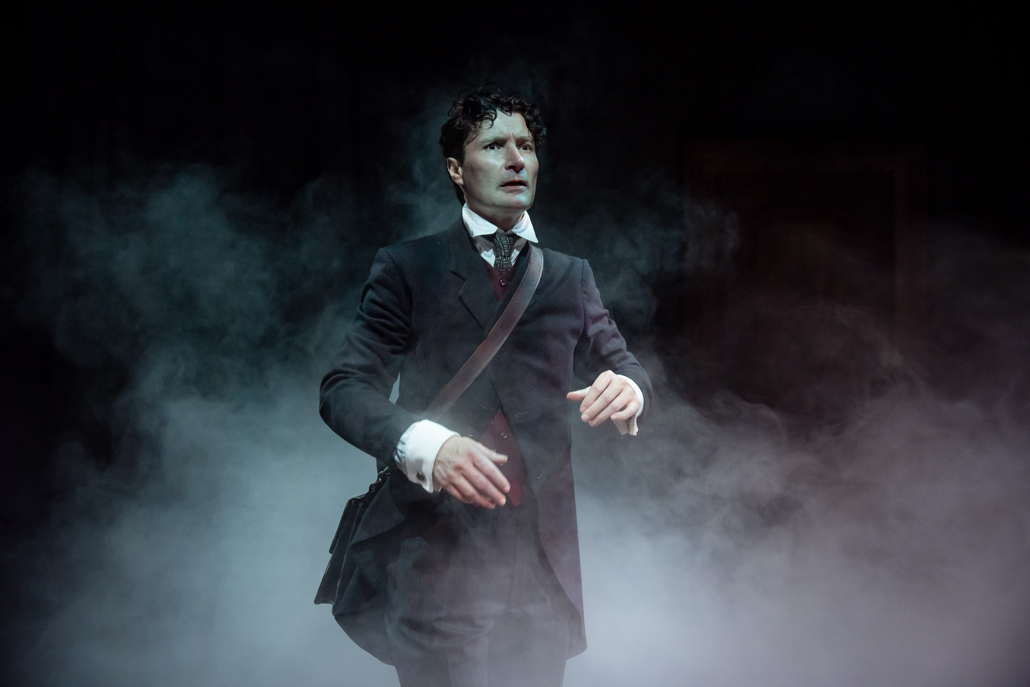 A man in a dark suit and tie stands in a dramatic cloud of smoke, looking ahead with a tense expression. He has curly hair and a shoulder bag, and appears to be on stage, illuminated by theatrical lighting.