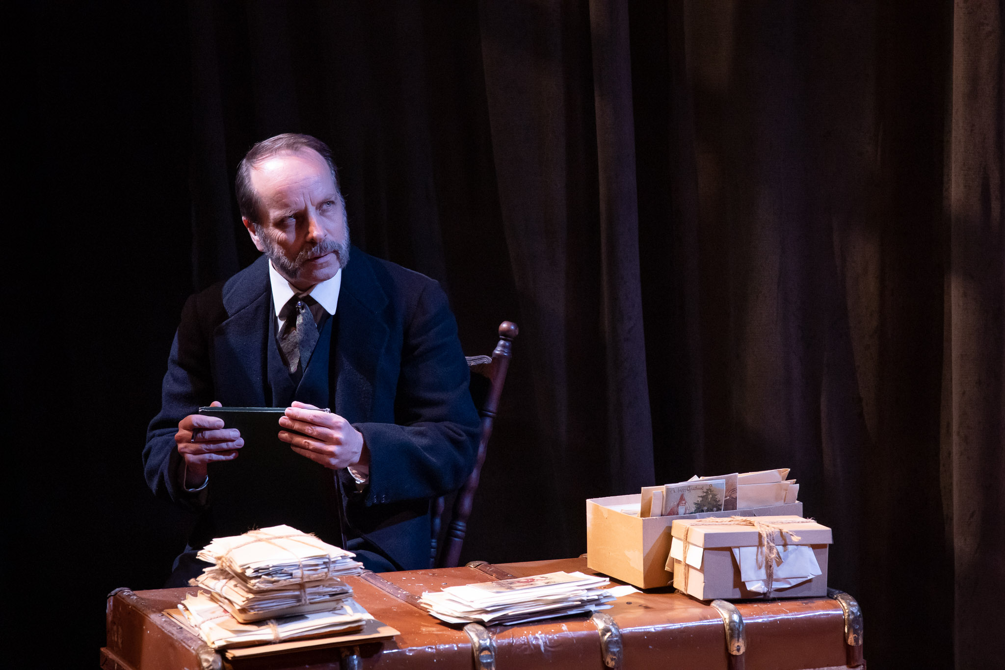 A man with a moustache, dressed in a suit, sits on a chair holding a tablet. He is next to a table with a stack of papers and boxes, set against a dark curtain backdrop, creating a dramatic atmosphere.