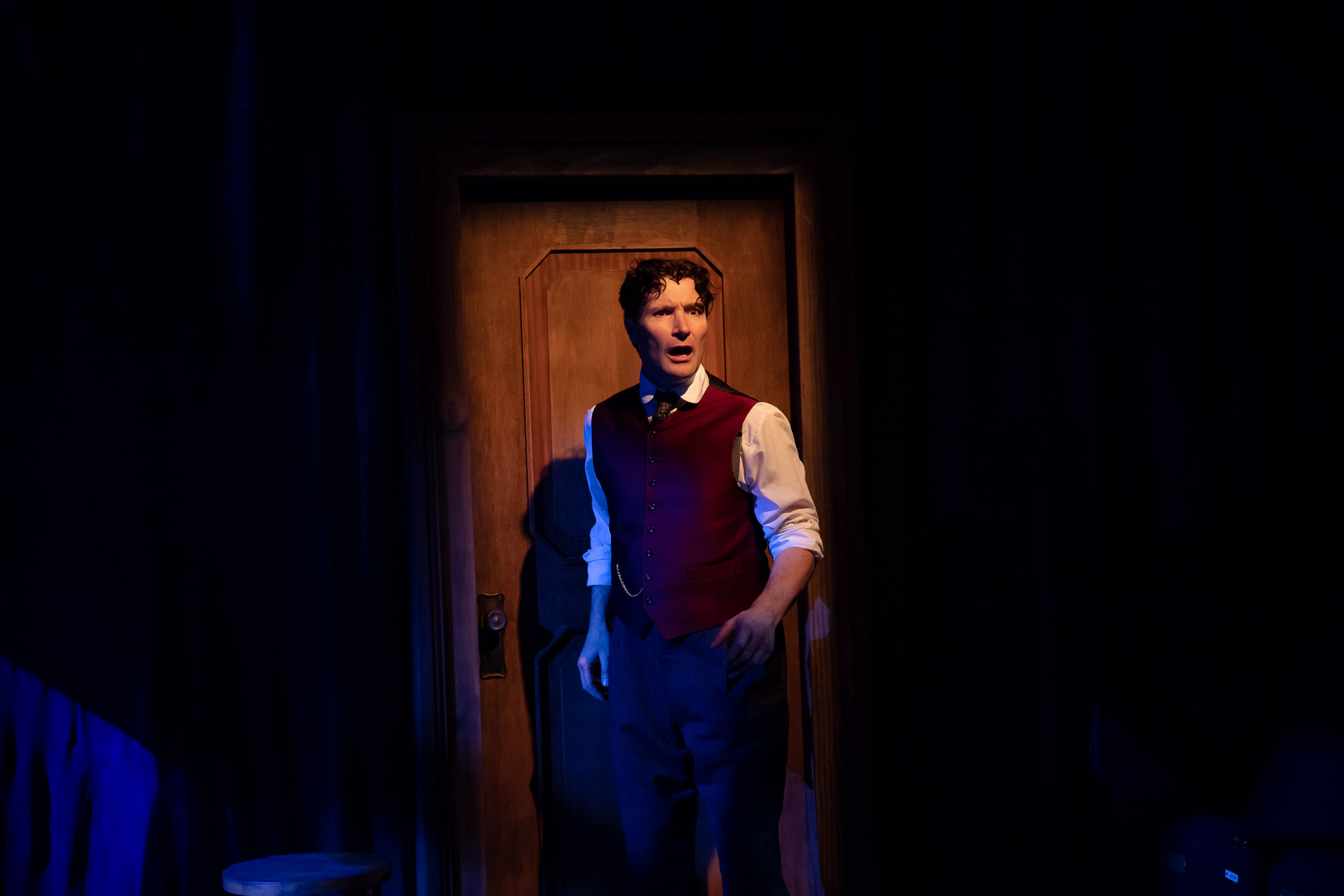 A man in a red waistcoat and white shirt stands in front of a wooden door on a dimly lit stage, illuminated by a spotlight, looking slightly to the side with a serious expression.