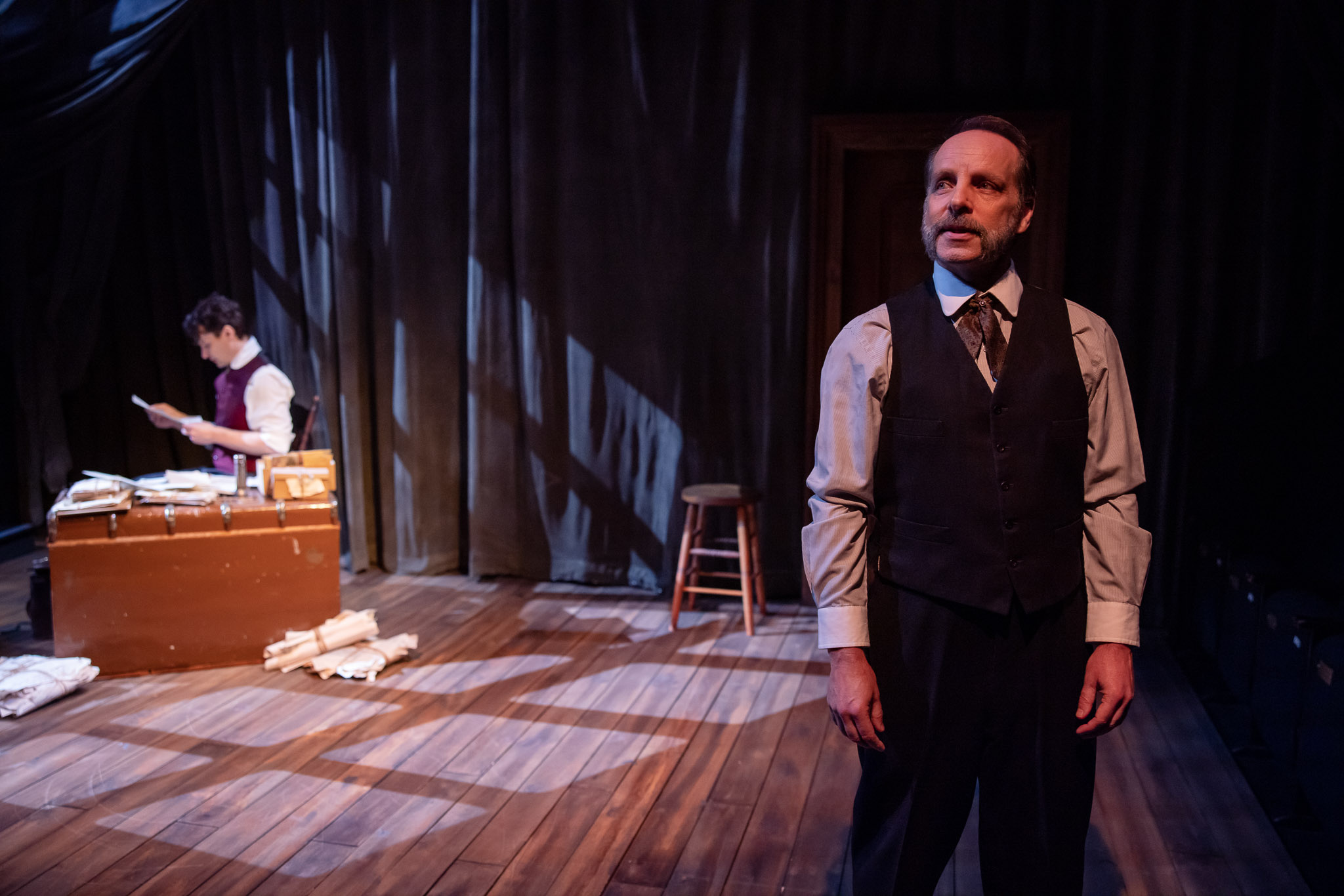 A man in a waistcoat stands in the foreground of a dimly lit stage, looking thoughtful. Behind him, another person reads papers at a cluttered desk. Shadows and dark curtains create a dramatic atmosphere.