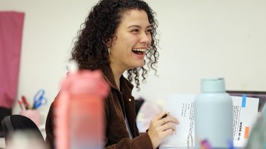 A young woman with curly hair smiles whilst sitting at a desk, holding papers and a pen. Various water bottles and colourful stationery items are on the desk in the foreground.