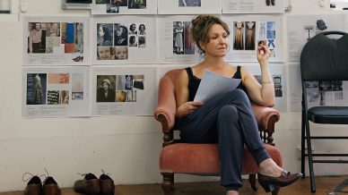 A woman sits on a pink armchair holding papers, appearing thoughtful. Behind her, mood boards and photos are pinned to the wall. Several pairs of shoes are lined up on the floor beside her. An empty chair is nearby.